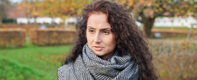 A woman with long curly hair and a gray scarf stands outdoors in a park, soaking in the autumn foliage.