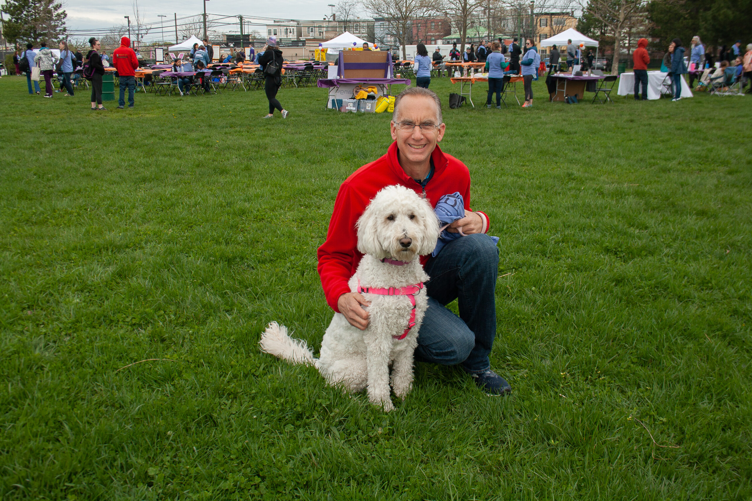 A man in a red jacket kneels beside his fluffy white dog, wearing a pink harness, on a grassy field at an outdoor gathering, debunking myths about man's best friend.