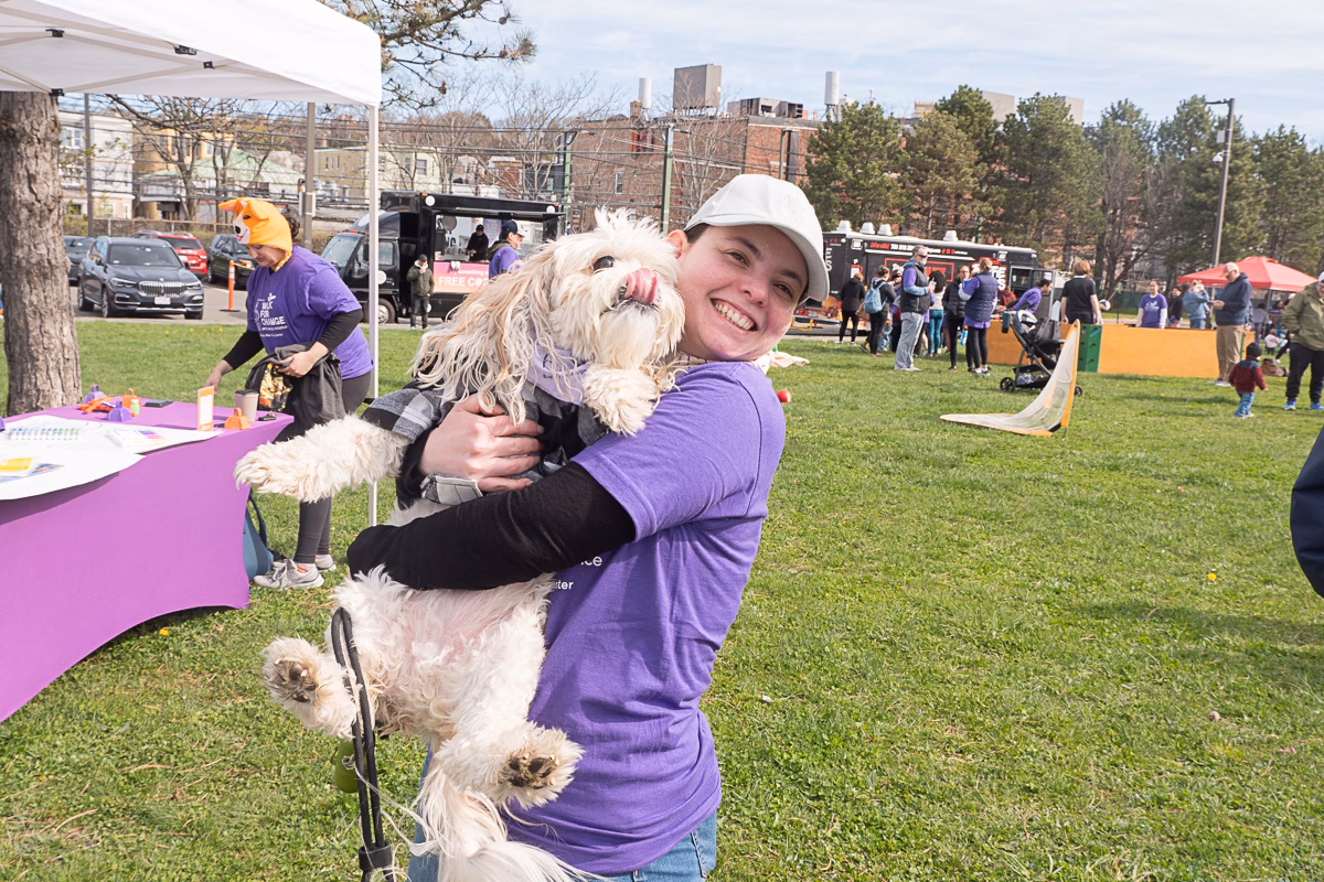 Smiling in a purple shirt, a person holds a fluffy dog at an outdoor event where myths blend into merry gatherings under tents with people in the background.