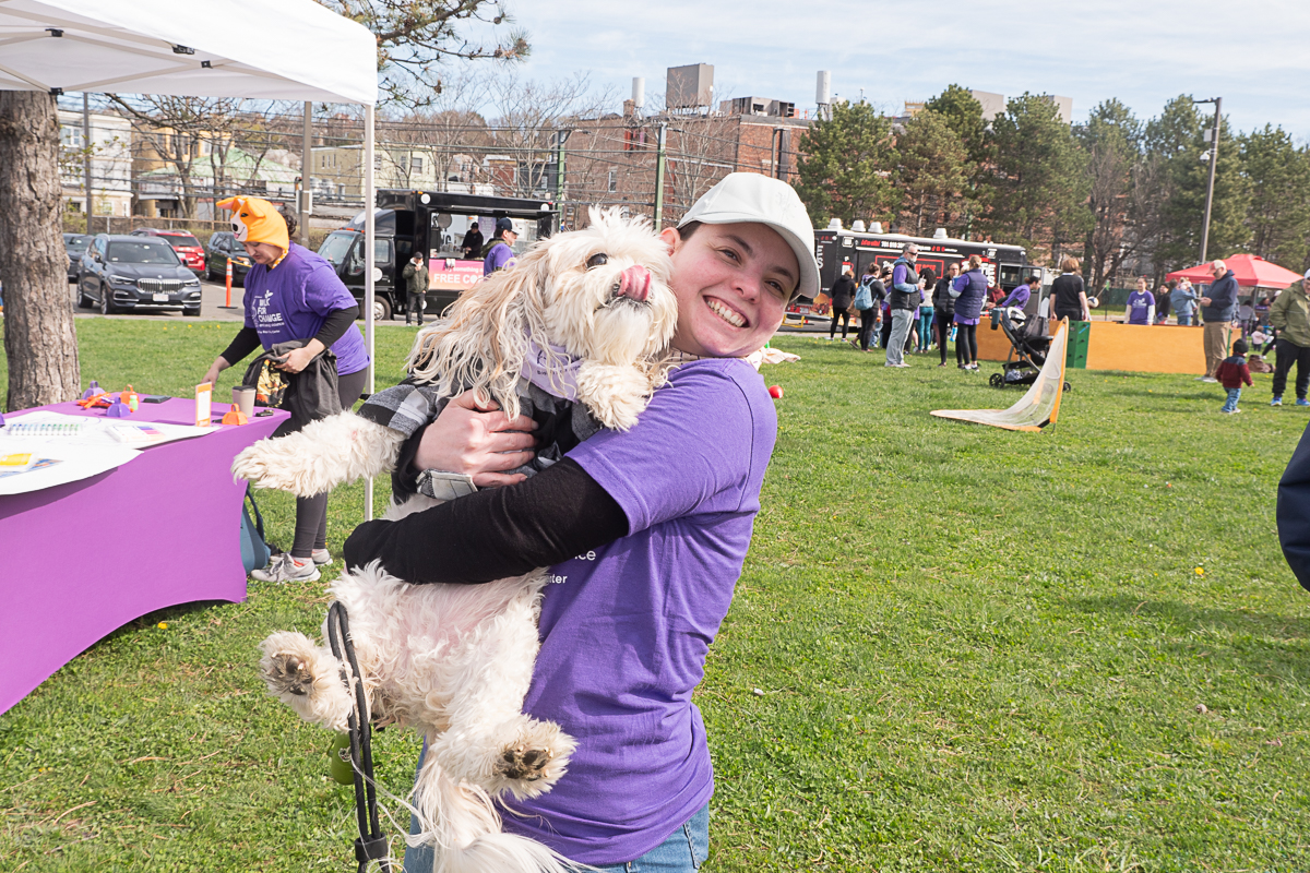 Smiling in a purple shirt, a person holds a fluffy dog at an outdoor event where myths blend into merry gatherings under tents with people in the background.