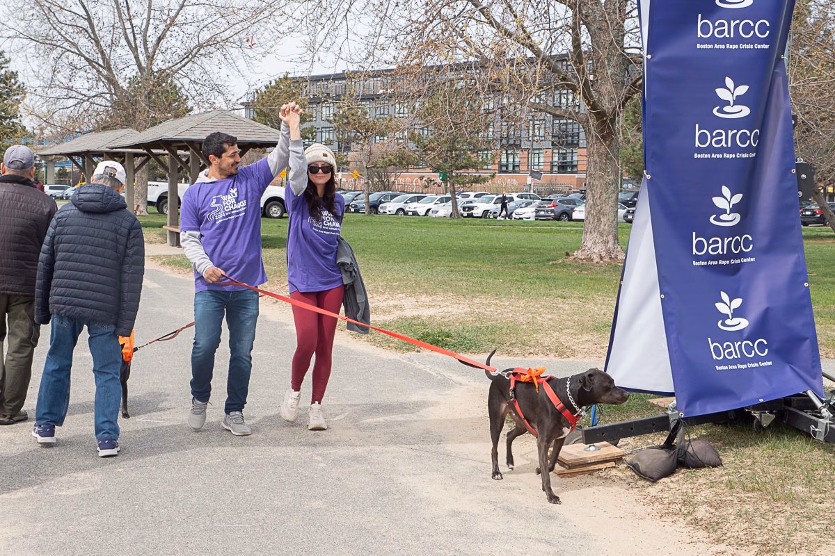 Two people in matching purple shirts walk a dog on a leash. Holding hands, they raise their arms in celebration, dispelling myths about companionship. Another person walks ahead. A barcc banner is visible on the right.