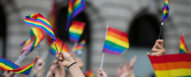 A crowd holds up rainbow flags at an outdoor event, celebrating Walk for Change with BARCC and sponsors, with blurred buildings in the background.