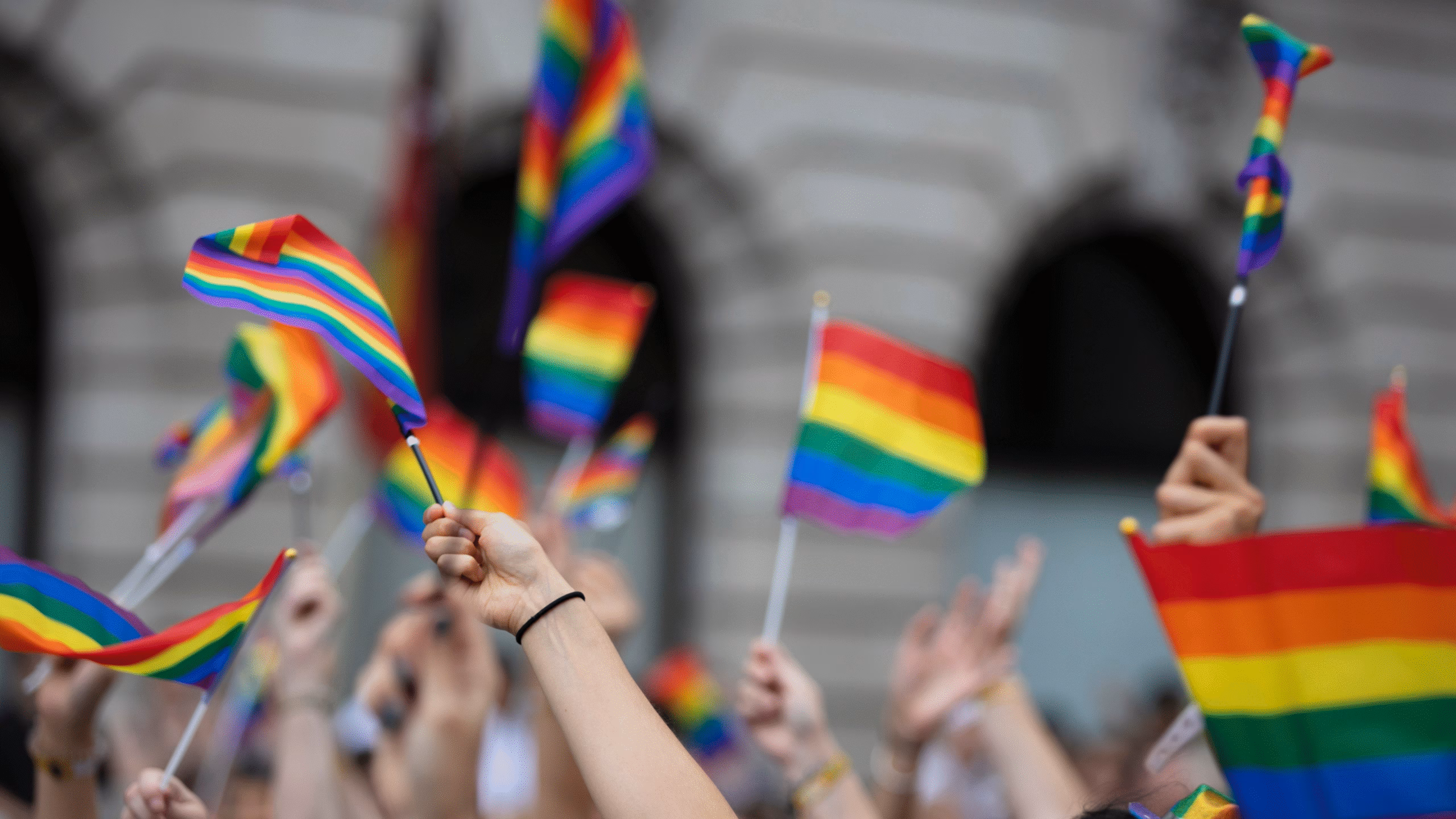 A crowd holds up rainbow flags at an outdoor event, celebrating Walk for Change with BARCC and sponsors, with blurred buildings in the background.