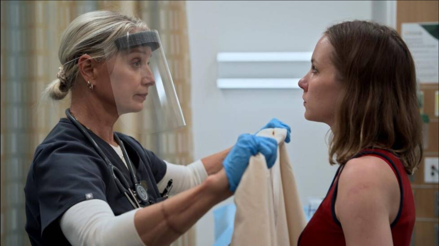 A healthcare worker in scrubs and a face shield holds up a gown for a woman in a sleeveless top, ensuring her consent inside a medical facility.