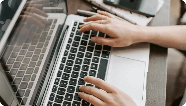 Manos escribiendo en el teclado de una computadora portátil, con un teléfono inteligente y una computadora portátil cerca.