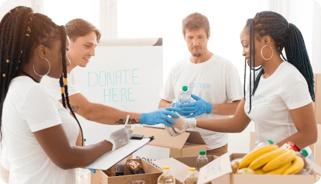 Four people in white shirts and gloves sort donations into boxes labeled "Donate Here," including bottled water and bananas.