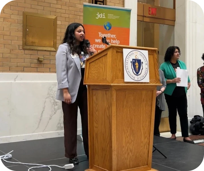A person speaks at a podium with a microphone. A banner in the background reads "Together, we can help create...". Two other individuals stand nearby.