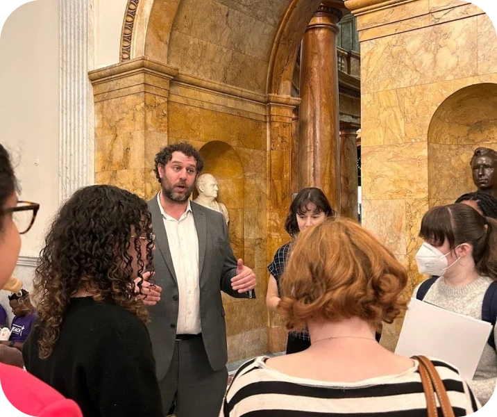 A man talks to a group of people in a building with marble columns and arches. Some listeners hold papers.