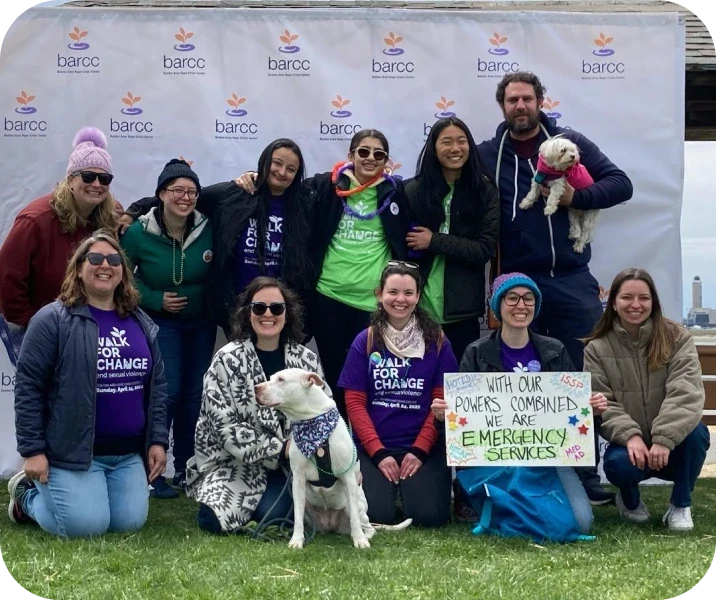 A group of people and two dogs pose in front of a "barcc" banner. One person holds a sign reading, "With our powers combined, we are emergency services.