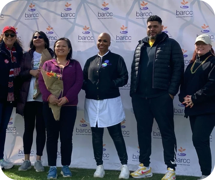 A group of six people posing in front of a backdrop with the BARCC logo. One person holds a bouquet of flowers.