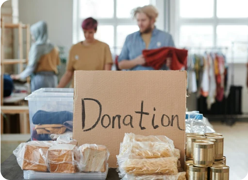 A donation table with food and clothing, featuring a cardboard sign labeled "Donation." In the background, two people sort through clothes.