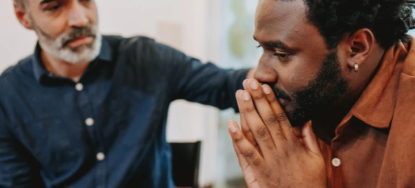 In a quiet Boston room, two men sit together. One man, looking concerned with hands over his mouth, is gently reassured by the other, whose supportive hand rests on his shoulder. They appear to be seeking comfort and perhaps considering the option to get help through available services.