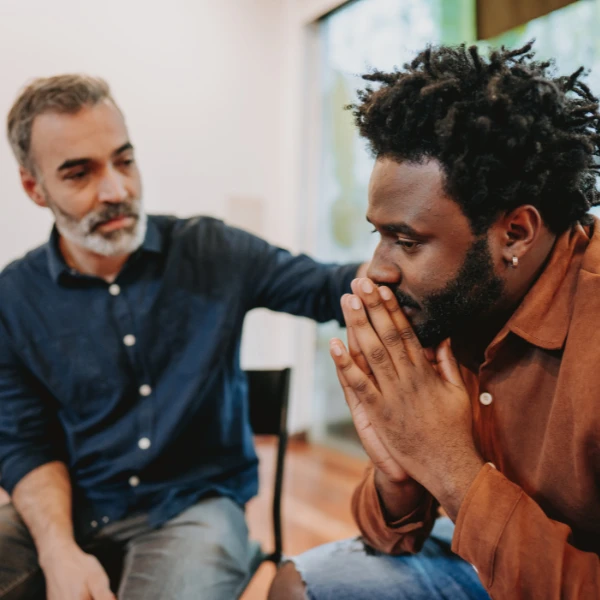 In a quiet Boston room, two men sit together. One man, looking concerned with hands over his mouth, is gently reassured by the other, whose supportive hand rests on his shoulder. They appear to be seeking comfort and perhaps considering the option to get help through available services.