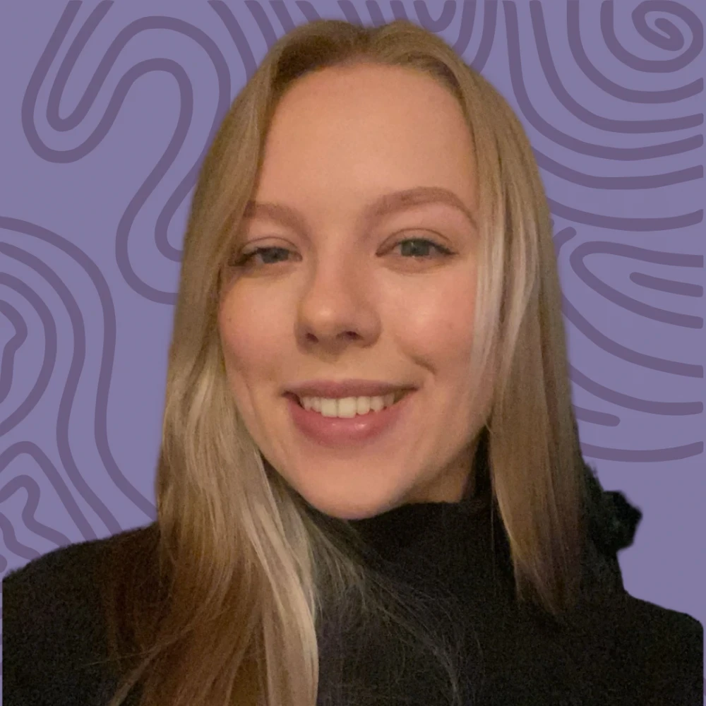 A smiling person with long blonde hair in a black top stands against a purple, wavy patterned background, embodying the warmth and support found at Boston's Rape Crisis Center.