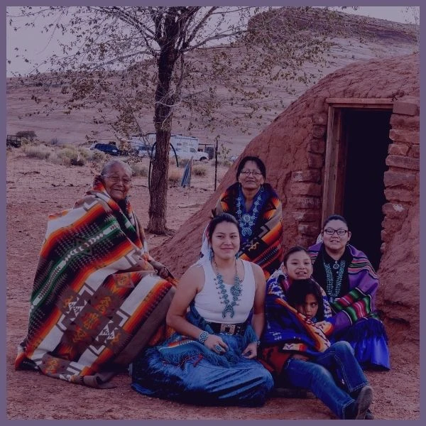 A group of people sit on the ground in front of a traditional clay structure, wrapped in colorful woven blankets. With the support of a Boston-based service, they find solace amid the desert landscape, under the sheltering tree.