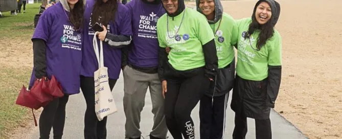 A group of people in raincoats and hats smiling at Boston's "Walk for Change" event, standing on a paved path near a grassy field. They are raising awareness and support for the vital services provided by a local rape crisis center.