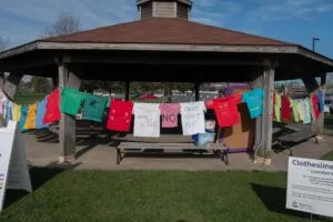 A Boston gazebo showcases colorful shirts with messages about breaking silence and educating against violence, urging the community to get involved as part of a public awareness display.