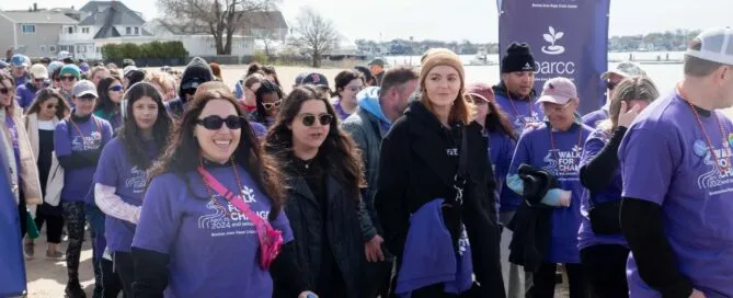A lively group in purple shirts gathers near the waterfront for an event, some sipping drinks and sporting sunglasses. Beneath a banner emblazoned with BARCC logos, they exude a vibrant community spirit.