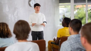 Classy Professional Real Estate Facebook Cover A person in a white sweater leads a Sexual Violence Prevention Training session, engaging with a group seated on chairs in a room adorned with windows and a clock.