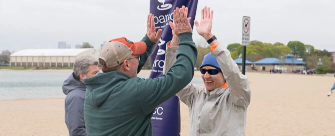Two men wearing jackets and hats give each other a high five on a sandy beach near a banner, with another person and lake in the background.