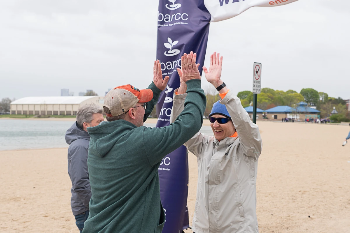 Two men wearing jackets and hats give each other a high five on a sandy beach near a banner, with another person and lake in the background.