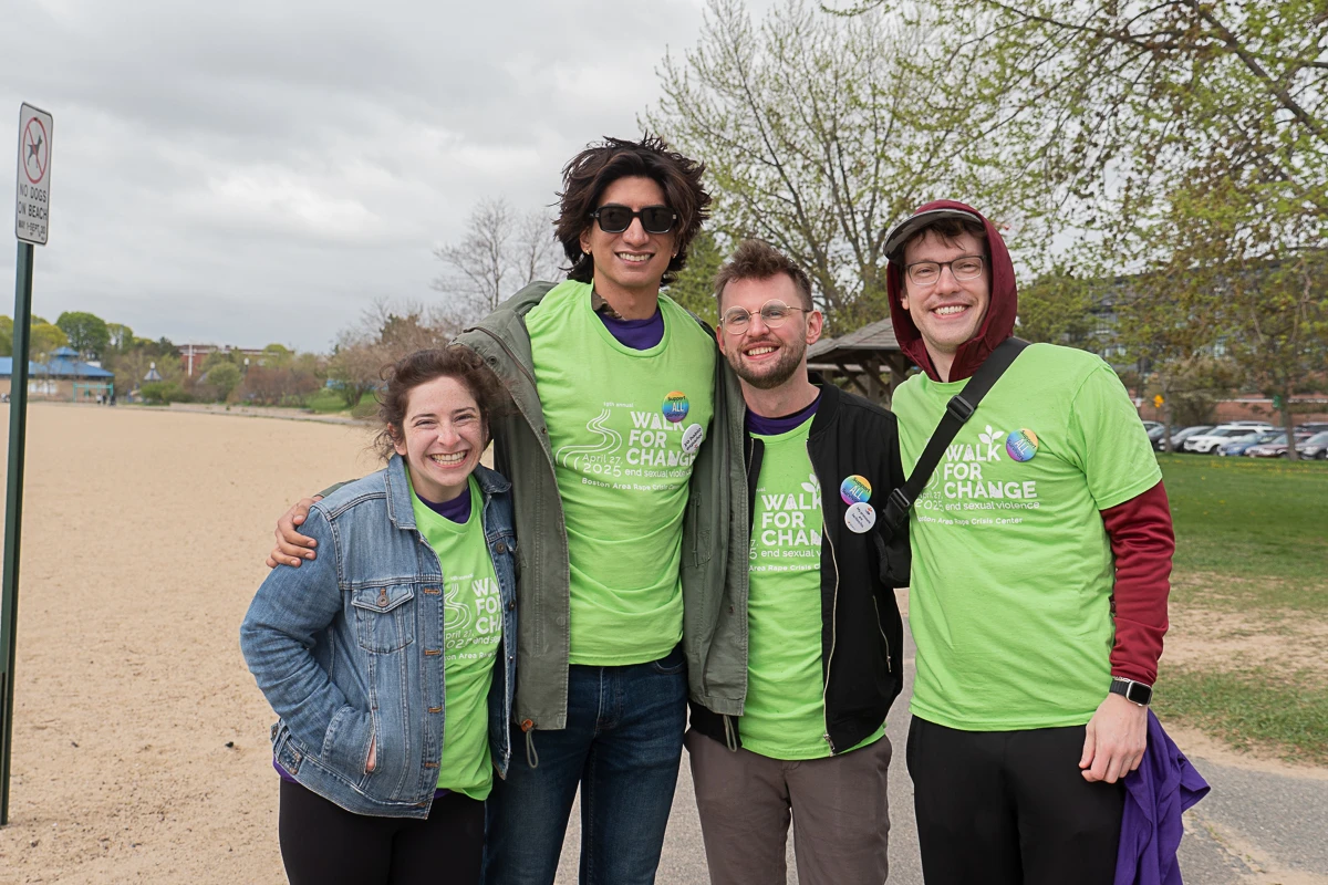 Four people wearing matching green "Walk for Change" shirts stand together outdoors, smiling at the camera near a sandy area with trees in the background.