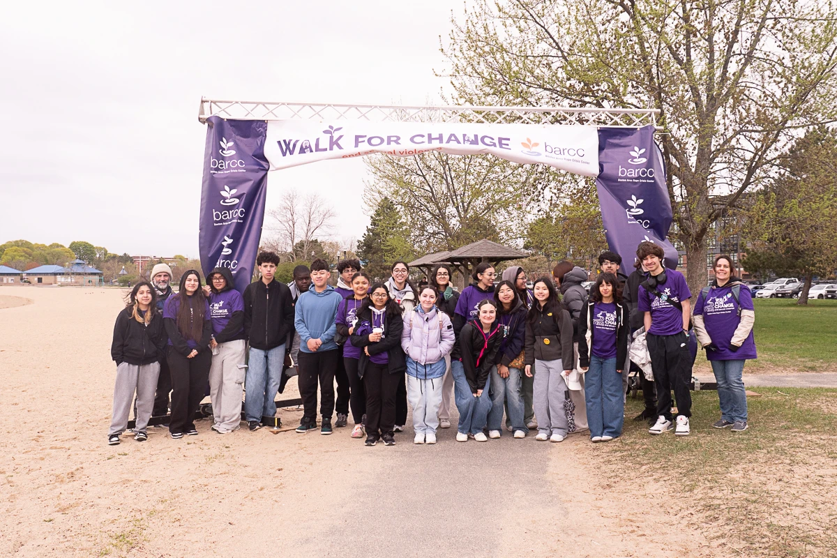 A group of people stands under a "Walk for Change" banner at an outdoor event, with trees and open space visible in the background.