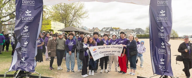 A group of people stand under a "Walk for Change" banner at a beach event organized by BARCC, holding signs in English and Spanish.