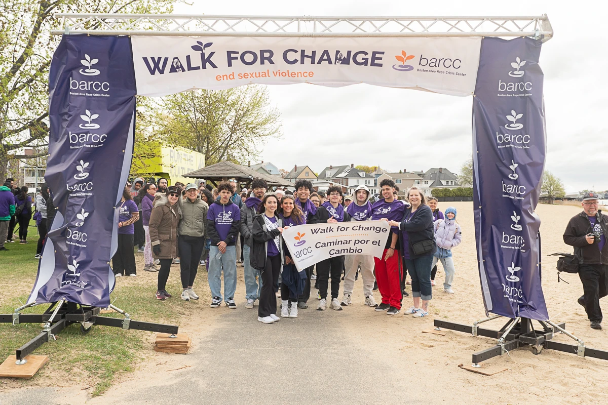 A group of people stand under a "Walk for Change" banner at a beach event organized by BARCC, holding signs in English and Spanish.
