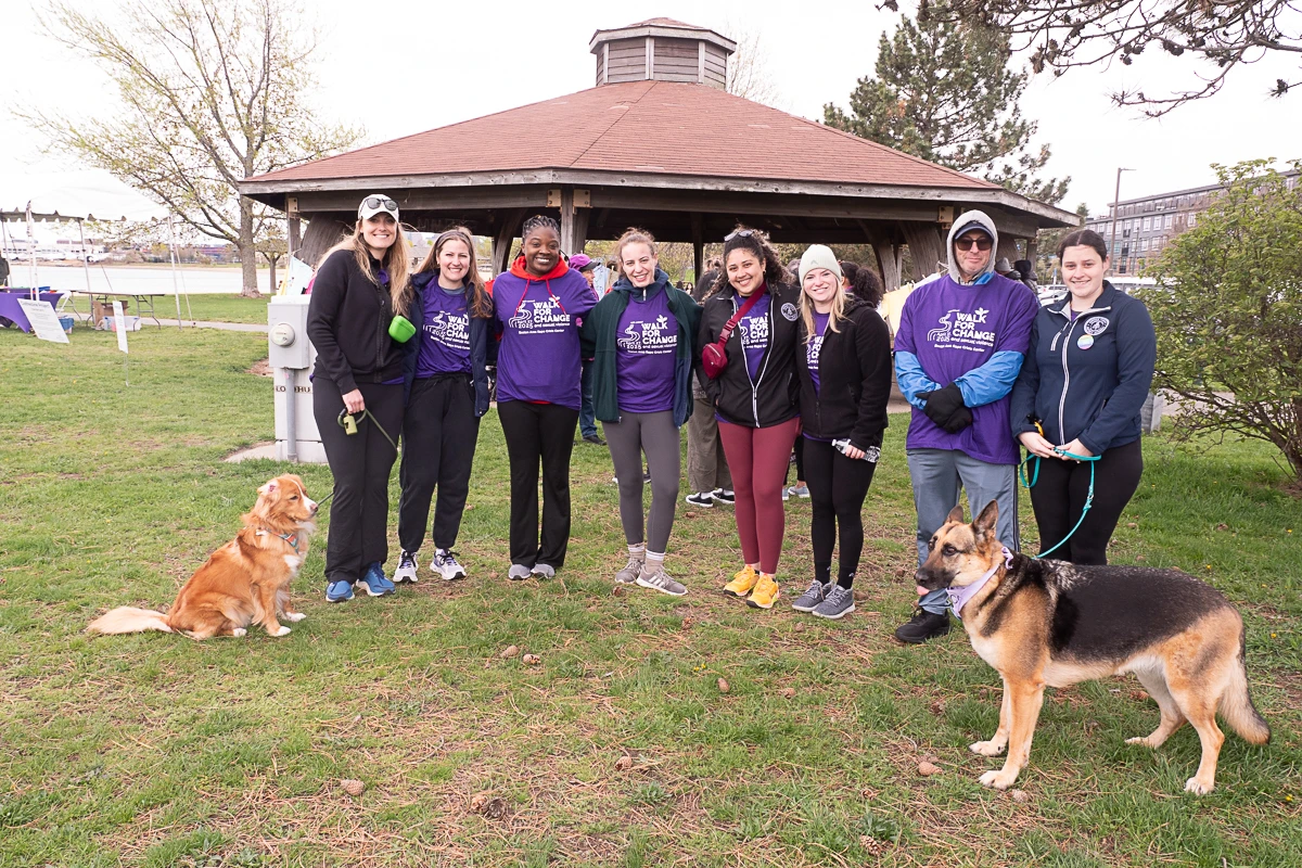 A group of nine people and two dogs stand together in front of a gazebo on a grassy area, posing for a photo during an outdoor event.