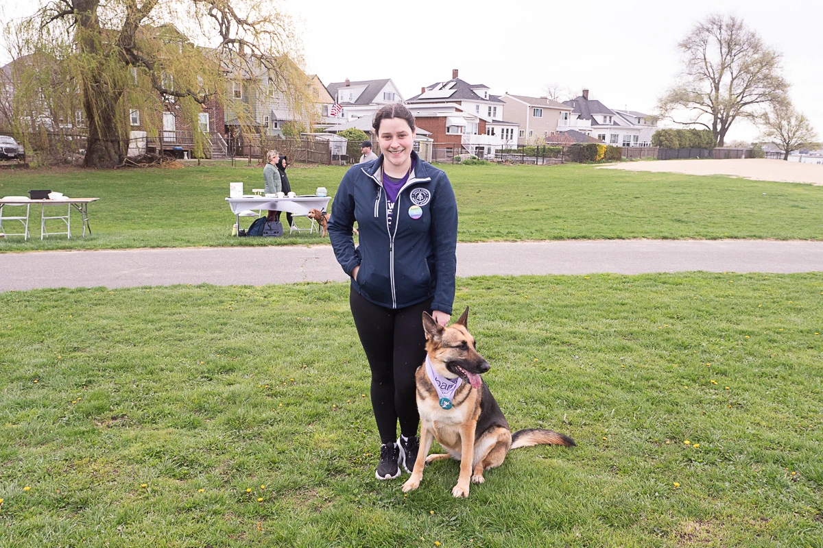A woman stands on grass next to a German Shepherd wearing a blue bandana, with houses and people at tables in the background.