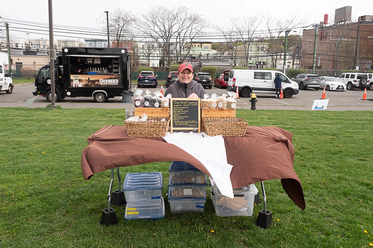 A person stands behind a table covered with baked goods and a menu board at an outdoor market, with plastic storage bins underneath and vehicles in the background.