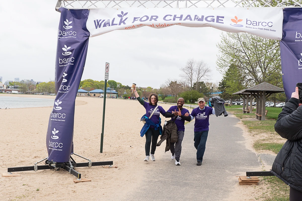 Three people in matching purple shirts celebrate as they cross the finish line at a “Walk for Change” event near a beach, with others and a photographer nearby.