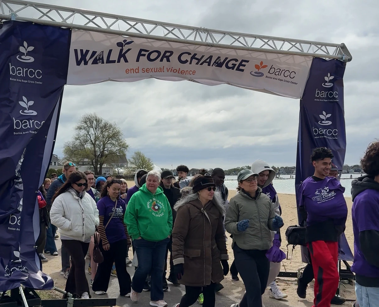 A group of people, including survivors, walk under a "Walk for Change: end sexual violence" banner on a sandy path during an organized SAAM event.