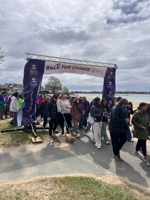 A group of people walk under a "Walk for Change" banner at an outdoor barcc event near a beach, some wearing jackets and walking dogs on a cloudy day as photos capture the moment.